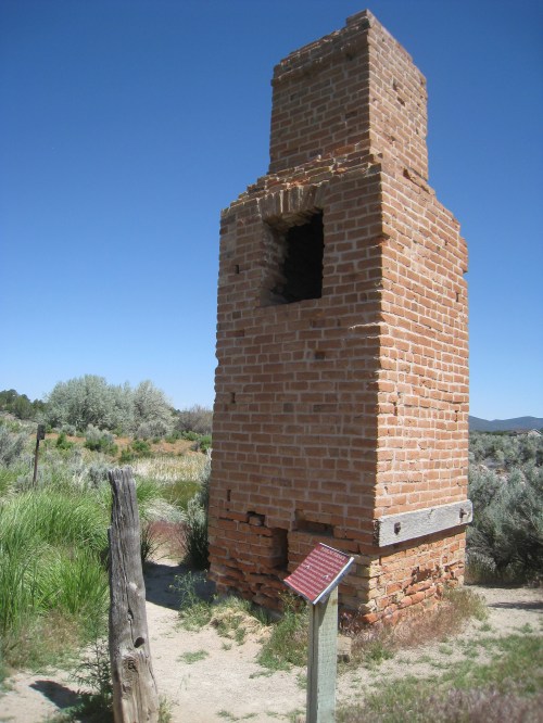 A weathered brick chimney stands alone in a grassy area, with a sign nearby and blue sky above.