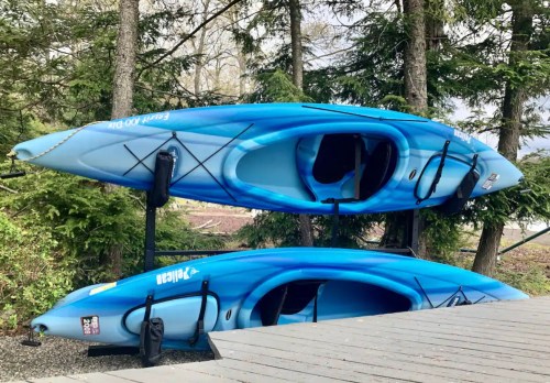 Two blue kayaks are stacked on a rack among green trees, with a wooden deck in the foreground.