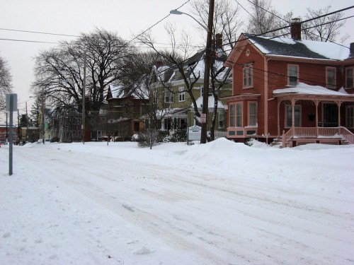 Snow-covered street lined with houses, trees bare, and power lines visible against a gray winter sky.