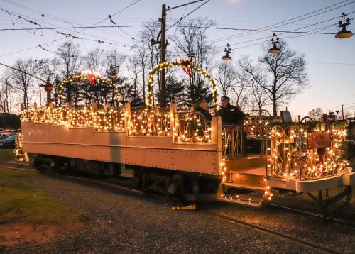 A decorated train covered in lights, parked on tracks at dusk, surrounded by trees and festive decorations.