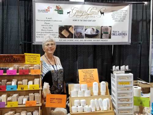 A vendor at a market showcasing handmade soaps and goat products, with colorful signs and a friendly smile.