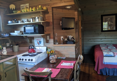 Cozy cabin interior featuring a kitchen with a stove, microwave, and dining table, plus a bed in the background.
