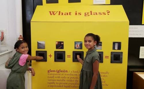Two children interact with a colorful exhibit titled "What is glass?" featuring black display panels.