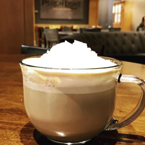 A close-up of a latte topped with whipped cream in a clear glass mug, set on a wooden table.