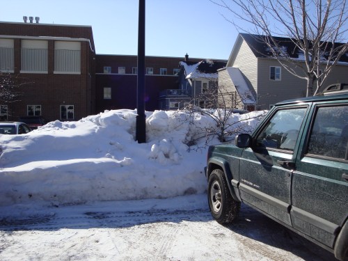 A snowy parking lot with a green vehicle parked beside a large snowbank and buildings in the background.