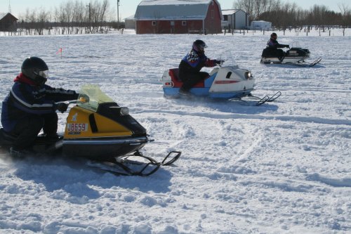 Three snowmobiles racing across a snowy field, with a red barn in the background and trees lining the horizon.