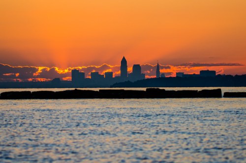 Silhouette of a city skyline against a vibrant orange sunset over calm waters.