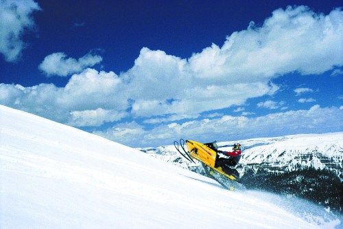 A person riding a yellow snowmobile on a snowy slope under a blue sky with fluffy clouds.