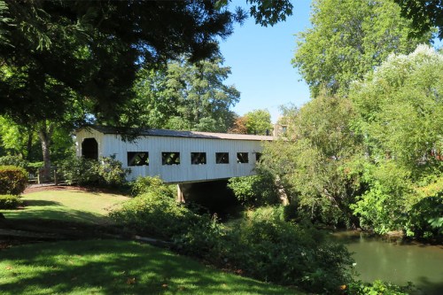 A white covered bridge spans a serene river, surrounded by lush greenery and trees under a clear blue sky.