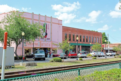 A colorful downtown scene featuring a pink building with a clock, shops, and parked cars along a tree-lined street.