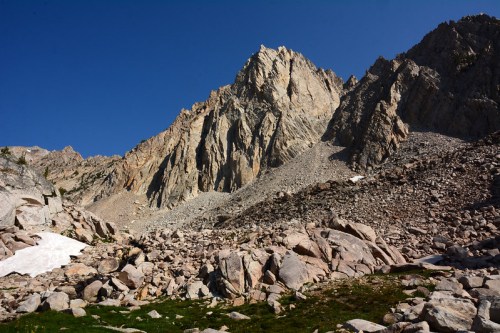 A rugged mountain landscape with steep rocky cliffs and scattered boulders under a clear blue sky.