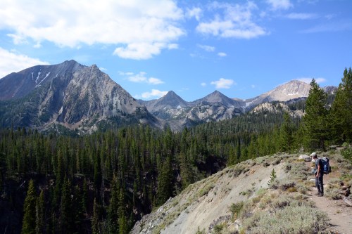 A hiker walks along a trail with mountains and pine trees under a blue sky with scattered clouds.