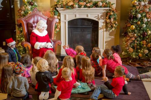 Mrs. Claus sits in a chair by a fireplace, surrounded by children eagerly listening to her story in a festive setting.