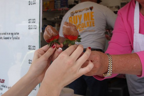 Two hands hold a chocolate-covered strawberry at a food stand, with a menu and a person in the background.