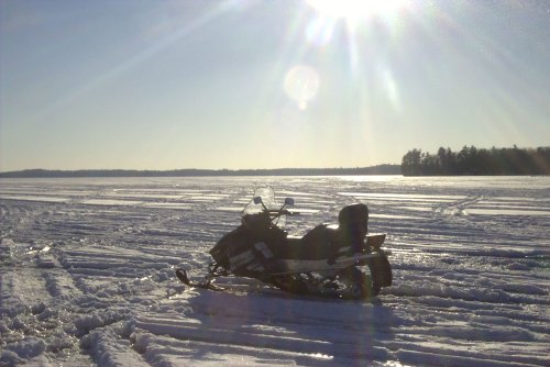 A snowmobile parked on a snowy landscape under a bright sun, with trees in the background and clear blue skies.