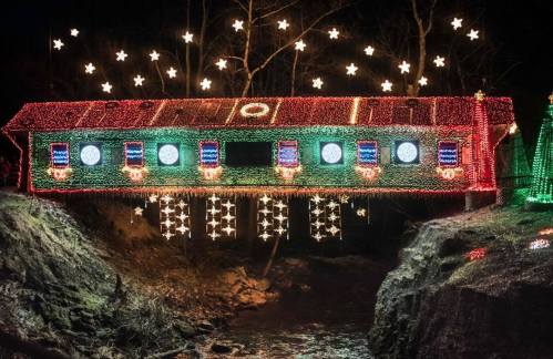 A brightly lit bridge decorated with colorful holiday lights and stars, spanning over a dark stream at night.