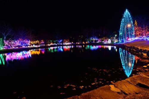 Colorful holiday lights reflect on a calm lake at night, with a glowing tree and festive decorations in the background.