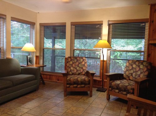 Cozy living space with two patterned chairs, a sofa, and large windows with blinds, illuminated by lamps.