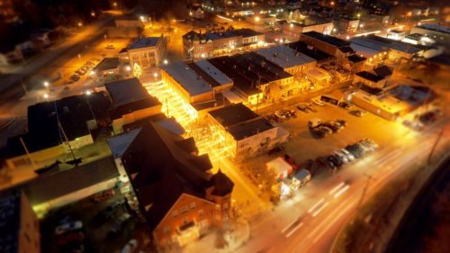 Aerial view of a brightly lit street at night, showcasing shops and buildings with warm lights and parked cars.