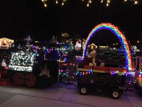 A festive train with colorful lights and a Santa figure, passing under a bright rainbow arch at night.