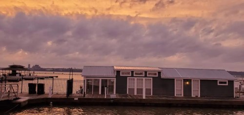 A serene waterfront scene featuring a houseboat against a colorful sunset sky with clouds.