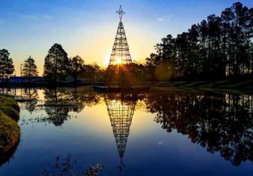 A metal Christmas tree silhouette reflects in a calm lake at sunset, surrounded by trees and a clear sky.