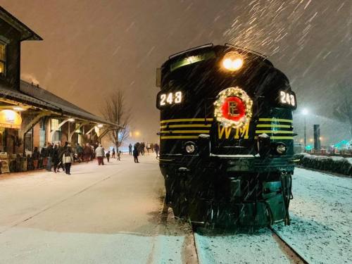 A vintage train adorned with a wreath stands on a snowy platform, with people gathered nearby in a festive atmosphere.