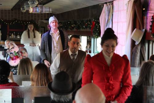 Actors perform in a festive train car, with guests seated at tables, decorated for the holiday season.