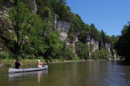Two people in a canoe on a calm river, surrounded by lush greenery and steep, rocky cliffs under a clear blue sky.