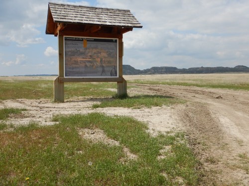 A weathered information sign stands beside a dirt path in a grassy landscape under a cloudy sky.