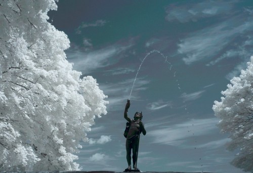 A statue of a figure holding a water spout, surrounded by trees and a cloudy sky, captured in infrared.