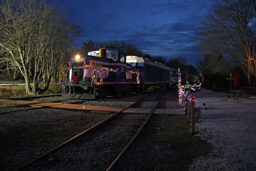 A decorated train with colorful lights parked on tracks at dusk, surrounded by bare trees and a peaceful setting.