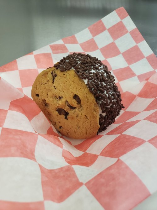 A cookie sandwich with chocolate chips, filled with ice cream and coated in chocolate sprinkles, on a red and white checkered paper.