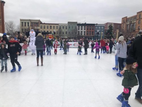 A busy outdoor ice skating rink with families and children skating, festive decorations, and a snowman character nearby.