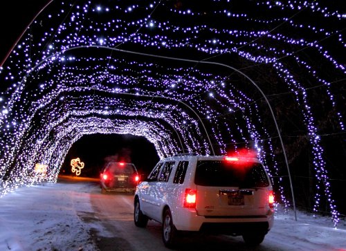 A white SUV drives through a tunnel of bright, twinkling lights at night, with snow on the ground.
