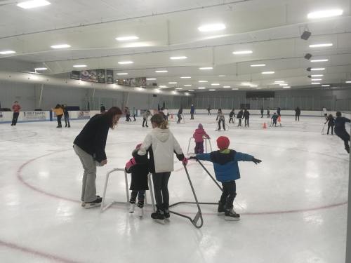 A busy ice skating rink with children and adults skating, some using support frames for balance.