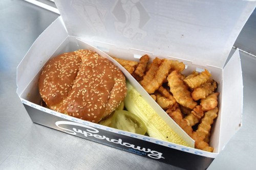A takeout box containing a sesame seed burger, crinkle-cut fries, and pickles.