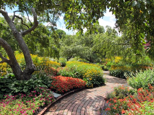 A vibrant garden filled with colorful flowers and lush greenery, featuring a winding brick path under a sunny sky.