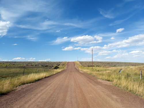 A dirt road stretches into the distance, flanked by grassy fields and under a blue sky with scattered clouds.