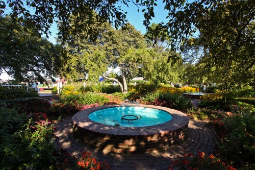 A circular stone fountain surrounded by vibrant flowers and lush greenery in a sunny garden setting.