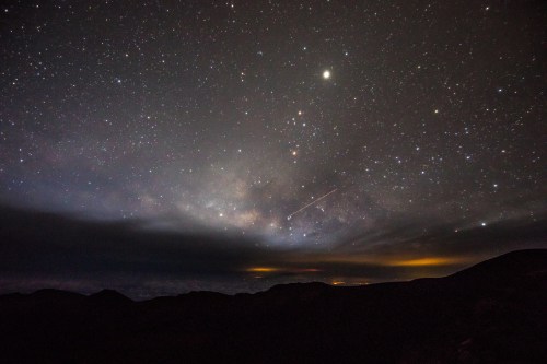 A starry night sky filled with stars, a faint Milky Way, and a shooting star over dark mountain silhouettes.