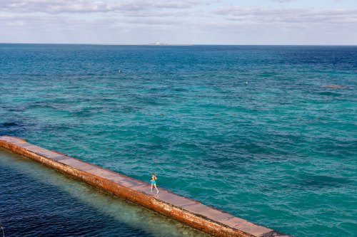A person walks along a weathered pier extending into turquoise waters under a partly cloudy sky.