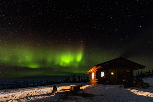 A cozy cabin under a starry sky, illuminated by vibrant green northern lights. Snow covers the ground.