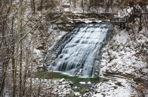 A snowy waterfall cascades down rocky cliffs, surrounded by bare trees and a serene winter landscape.