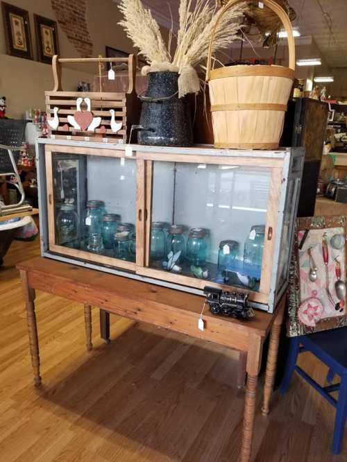 A vintage display cabinet with glass doors, showcasing jars, on a wooden table with various decorative items around it.