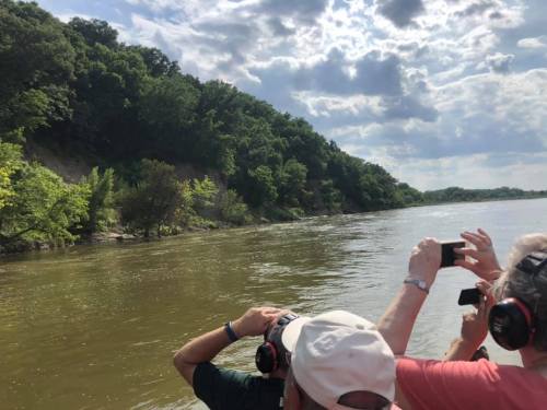 People wearing headphones take photos of a riverbank surrounded by lush greenery under a cloudy sky.