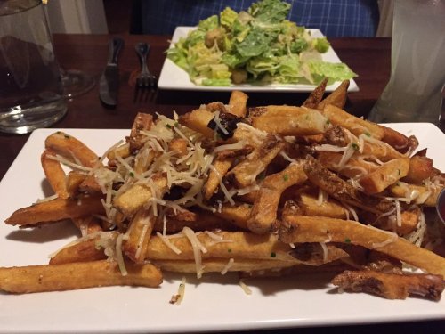 A plate of golden fries topped with cheese and herbs, served alongside a Caesar salad in the background.