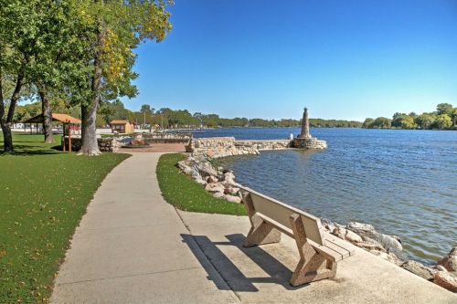 A serene lakeside view with a path, bench, and stone pier under a clear blue sky and lush green trees.