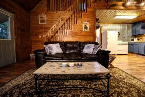 Cozy cabin interior featuring a leather sofa, wooden walls, a coffee table, and a staircase in the background.