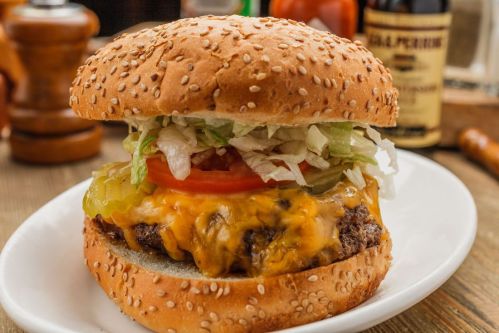 A close-up of a sesame seed burger with a beef patty, melted cheese, lettuce, tomato, and pickles on a white plate.